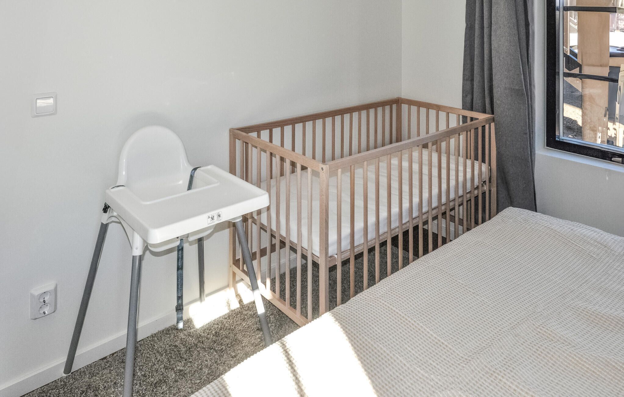 A baby crib with a wooden frame and a white mattress, a white baby high chair with a tray, and a window with grey curtains.