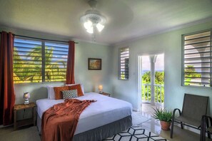 Une chambre à coucher comprenant un lit, une table de chevet, une lampe, une fenêtre avec des rideaux, un ventilateur de plafond et une vue sur la verdure à l’extérieur.