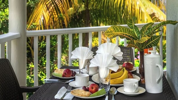 Mesa dispuesta con alimentos para el desayuno, una planta y una jarra blanca en un balcón con vistas a palmeras.
