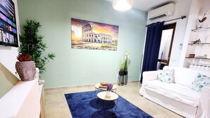A living room with a blue rug, a white sofa, a small round table, a potted plant, and a painting of the Colosseum. - Villetta Alessandra Ostia Antica 10 mins from beach (Lazio)