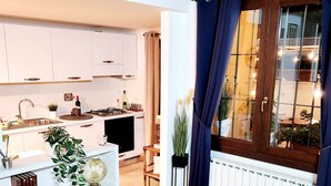 A modern kitchen with white cabinets, a stove, and a sink. There is a window with blue curtains and a view of the outside. - Villetta Alessandra Ostia Antica 10 mins from beach (Lazio)