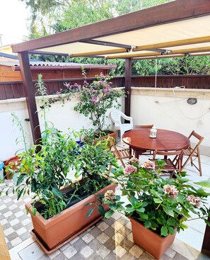Un patio con una mesa y sillas de madera, rodeado de plantas en macetas y una pérgola con dosel color beige.