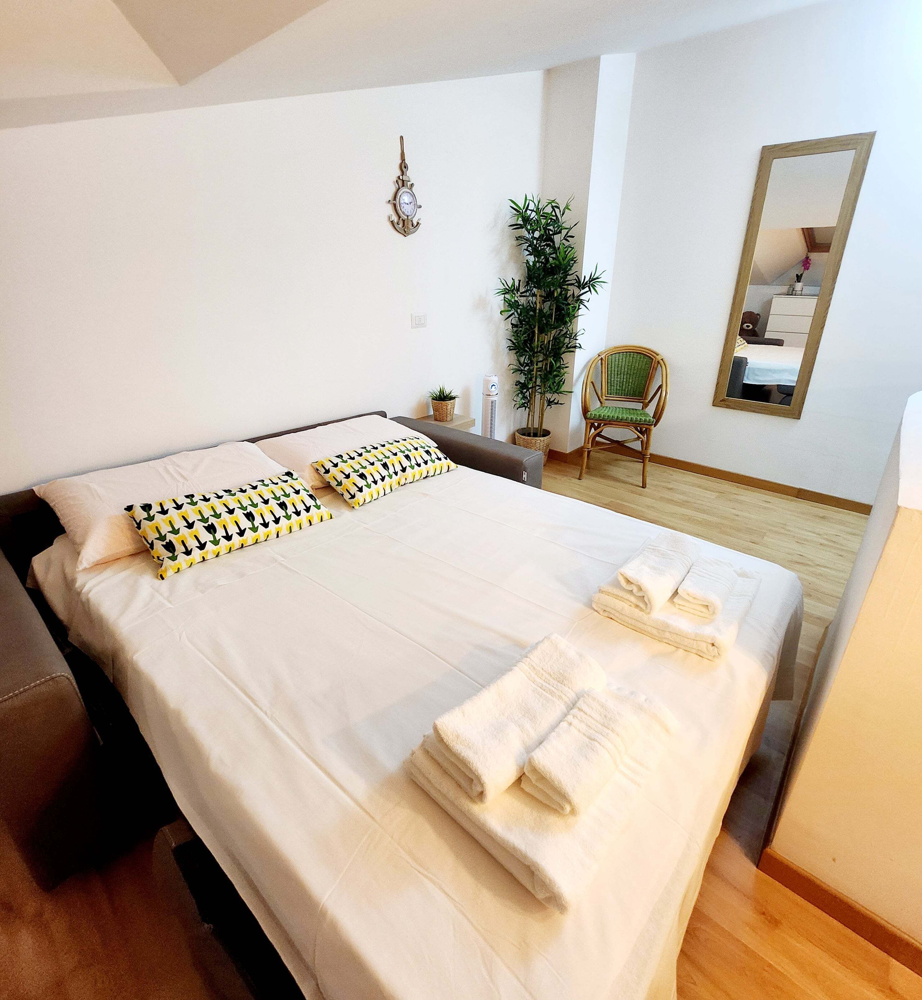 Un lit bien fait, avec du linge de lit blanc et des coussins à motifs, une table de chevet avec une plante, une chaise et un miroir au mur.