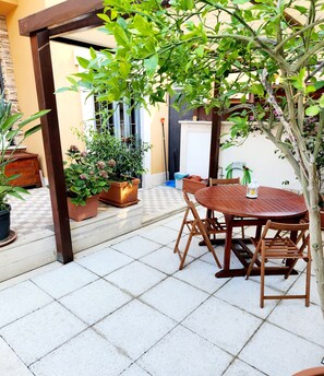 Un patio con mesa y sillas de madera, rodeado de plantas en macetas y una pérgola.