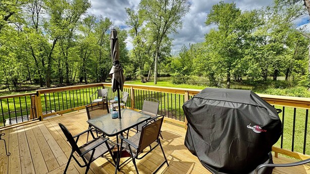 Una terraza de madera con una mesa, sillas, una funda para parrilla y vistas a un lago rodeado de árboles.