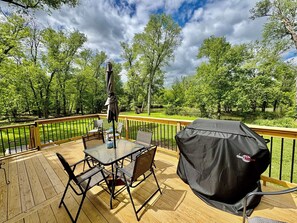 Una terraza de madera con una mesa, sillas, una funda para parrilla y vistas a un lago rodeado de árboles.