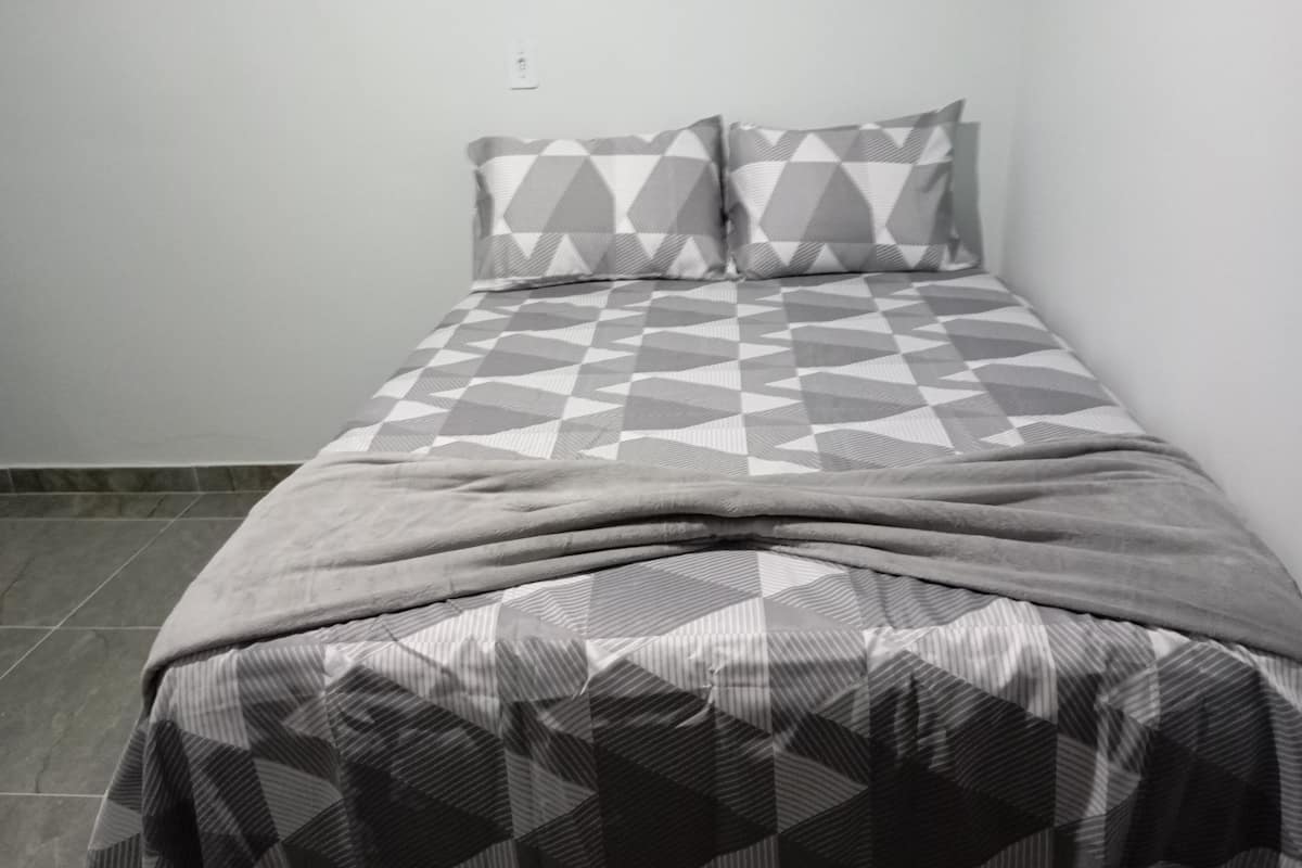 A bed with a geometric patterned bedspread and pillows, a plain wall, and a tiled floor.