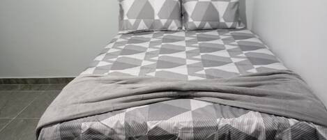 Un lit avec une couvre-lit et des oreillers aux motifs géométriques, un mur uni et un sol carrelé.