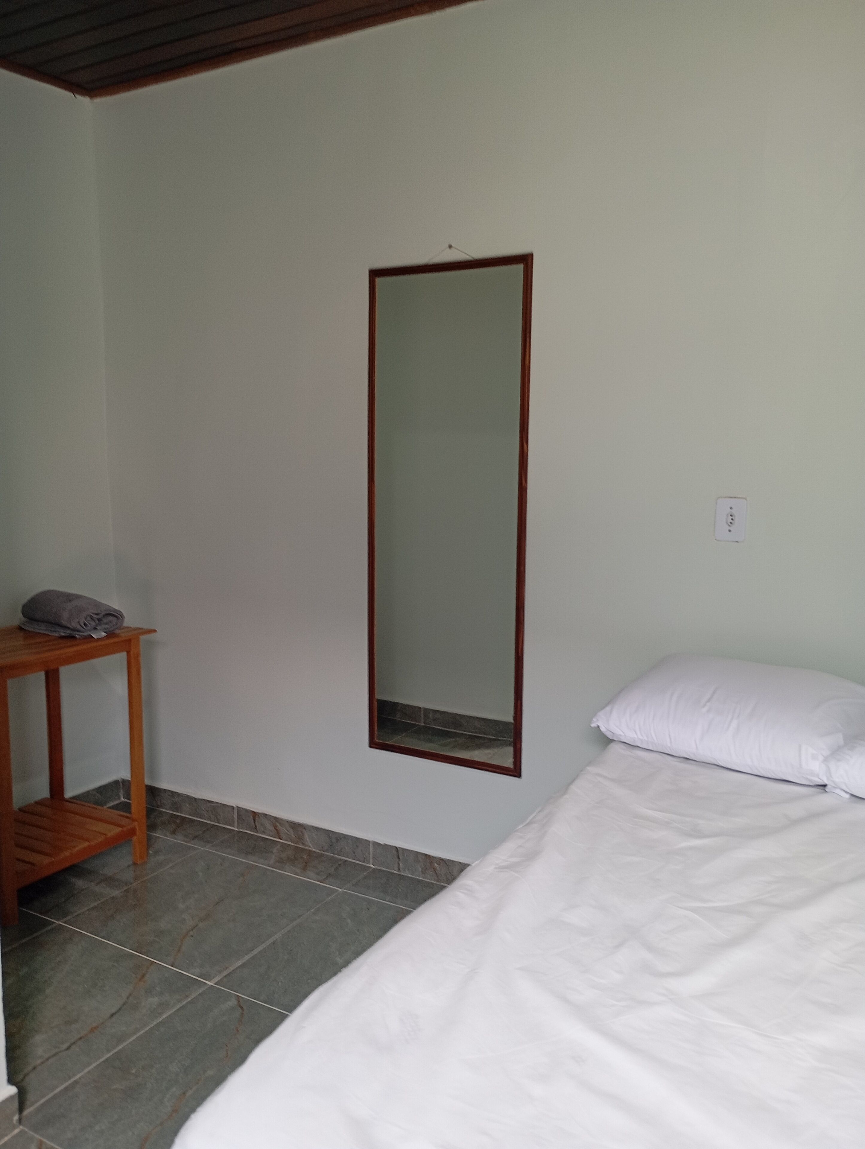 A bedroom with a bed, a wooden nightstand, a mirror, and a wall socket.