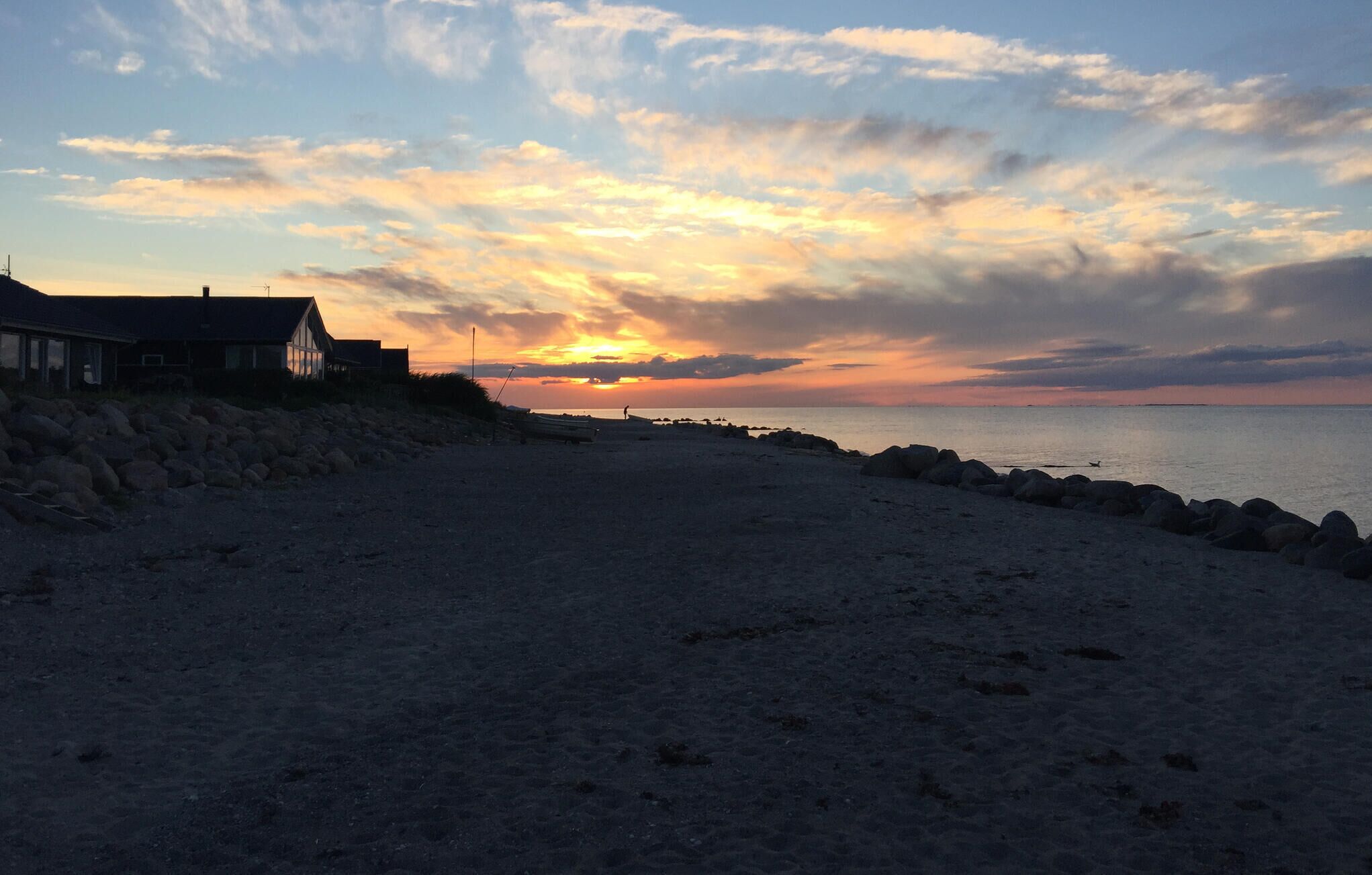 A beach with a house on the left, a rocky shoreline, and a sunset in the background.