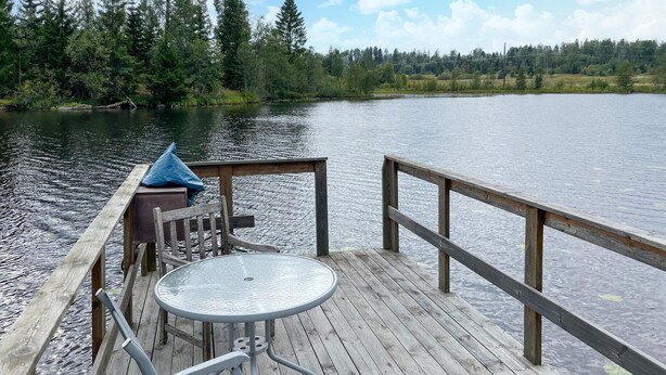A wooden dock with a metal table and chairs, overlooking a calm lake with trees in the background.