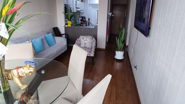 A modern living room with a glass coffee table, a sofa with blue cushions, and a dining area with a glass table and chairs. A kitchen is visible in the background. - Cozy apartment (Pueblo libre)