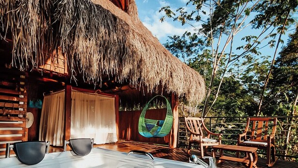 A wooden deck with a hot tub, wooden chairs, and a thatched-roof structure. - Refugio Monte Oscuro (La Dorada)
