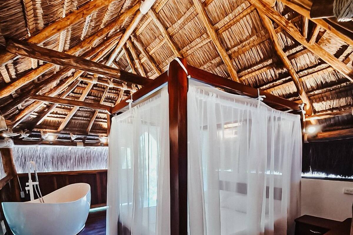 A room with a thatched roof, a white canopy bed, a large white bathtub, and wooden flooring.