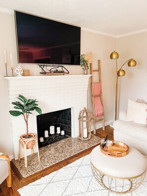 A living room with a white brick fireplace, a flat-screen TV mounted above it, a potted plant, and a round ottoman with a tray.