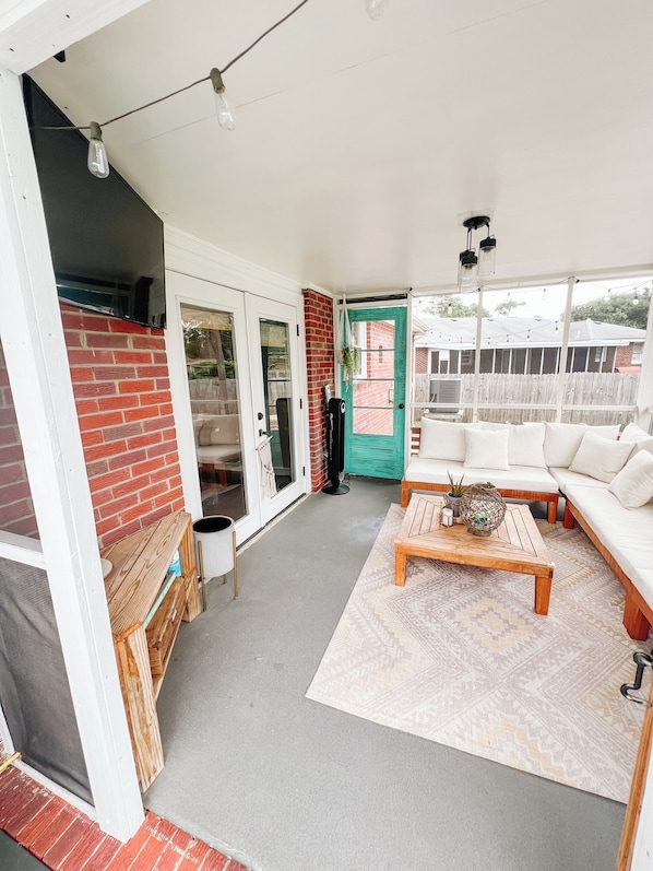 A spacious living area with a white sofa, a wooden coffee table, and a patterned rug. There is a brick wall and a green door leading outside.