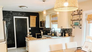 Una cocina con isla central, muebles blancos, una pizarra con escritura a tiza y una lámpara colgante.