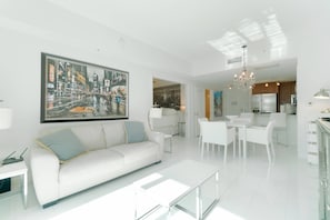 Uma sala de estar moderna com sofá branco, área de jantar com cadeiras brancas, cozinha com eletrodomésticos de aço inoxidável e uma grande pintura de paisagem urbana na parede.