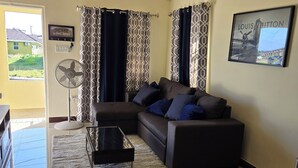 Un salon avec un canapé d’angle, une table basse en verre, des rideaux à motifs, un ventilateur et un tableau encadré accroché au mur.