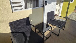 Un espace extérieur aménagé avec deux chaises noires et une petite table, une porte blanche et une fenêtre avec des stores.