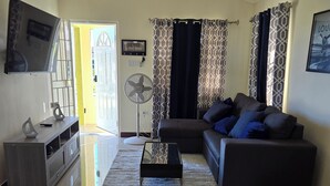 Un salon avec un canapé, une table basse, un téléviseur fixé au mur, un ventilateur et un rideau à motifs.