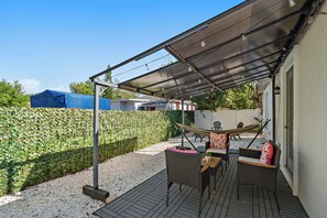 Een overdekte terras met een tafel, stoelen, een hangmat en een haag op de achtergrond.