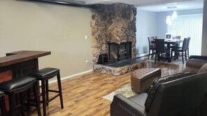 Una sala de estar con chimenea de piedra, piso de madera, una zona de bar con taburetes y un comedor con sillas y mesa.