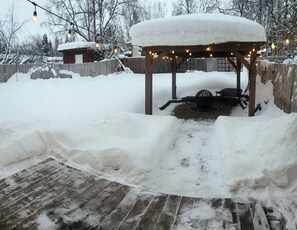 A snow-covered outdoor area with a wooden gazebo, string lights, and a fire pit.