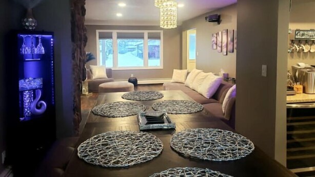 A modern living room with a large wooden table, multiple poufs, and a built-in entertainment unit with a screen.