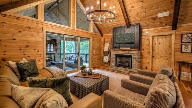 A cozy log cabin interior with a fireplace, comfortable seating, and a large window overlooking a deck. - The Retreat at Laurel Creek- Screened Porch, Hot Tub, Outdoor Fireplace (Blue Ridge)