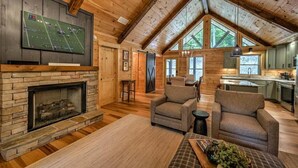 A spacious living room with a stone fireplace, comfortable seating, and a large TV mounted above it. - The Retreat at Laurel Creek- Screened Porch, Hot Tub, Outdoor Fireplace (Blue Ridge)