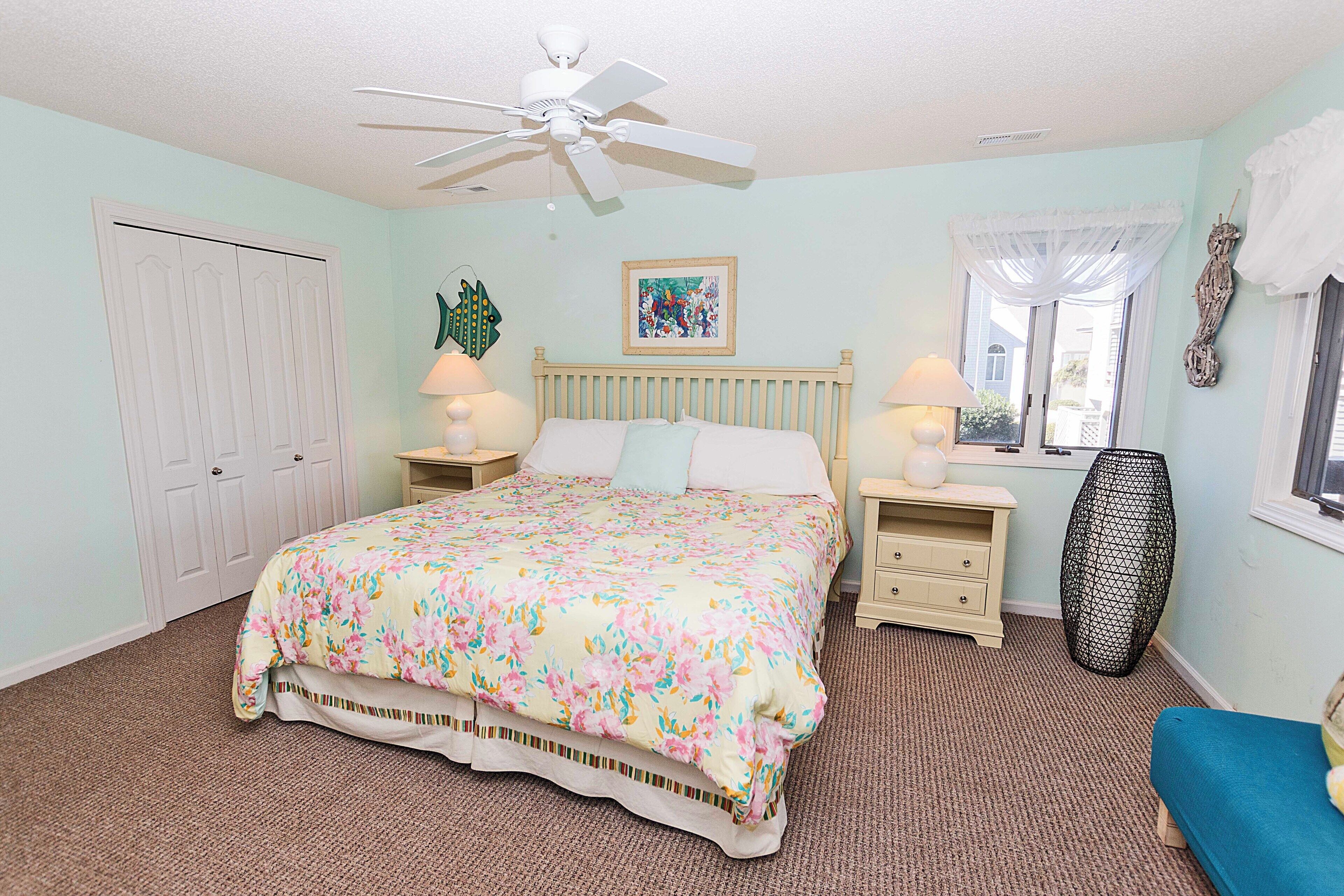 A bedroom with a bed, two bedside tables, a ceiling fan, a window with curtains, and a door.