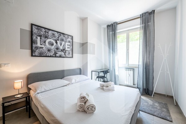 A hotel room with a bed, a bedside table with a lamp, a framed picture on the wall, a small table with chairs, and a window with curtains.