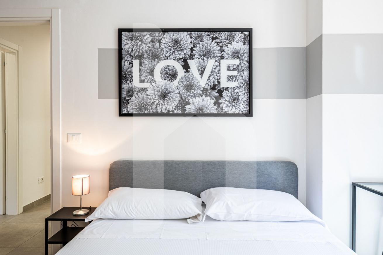 A bedroom with a bed, white pillows, a bedside lamp, and a framed picture on the wall.
