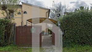 Une entrée fermée par un portail mène à un bâtiment, un chemin de gravier y conduit, et l’ensemble est entouré de verdure et d’une clôture en bois.