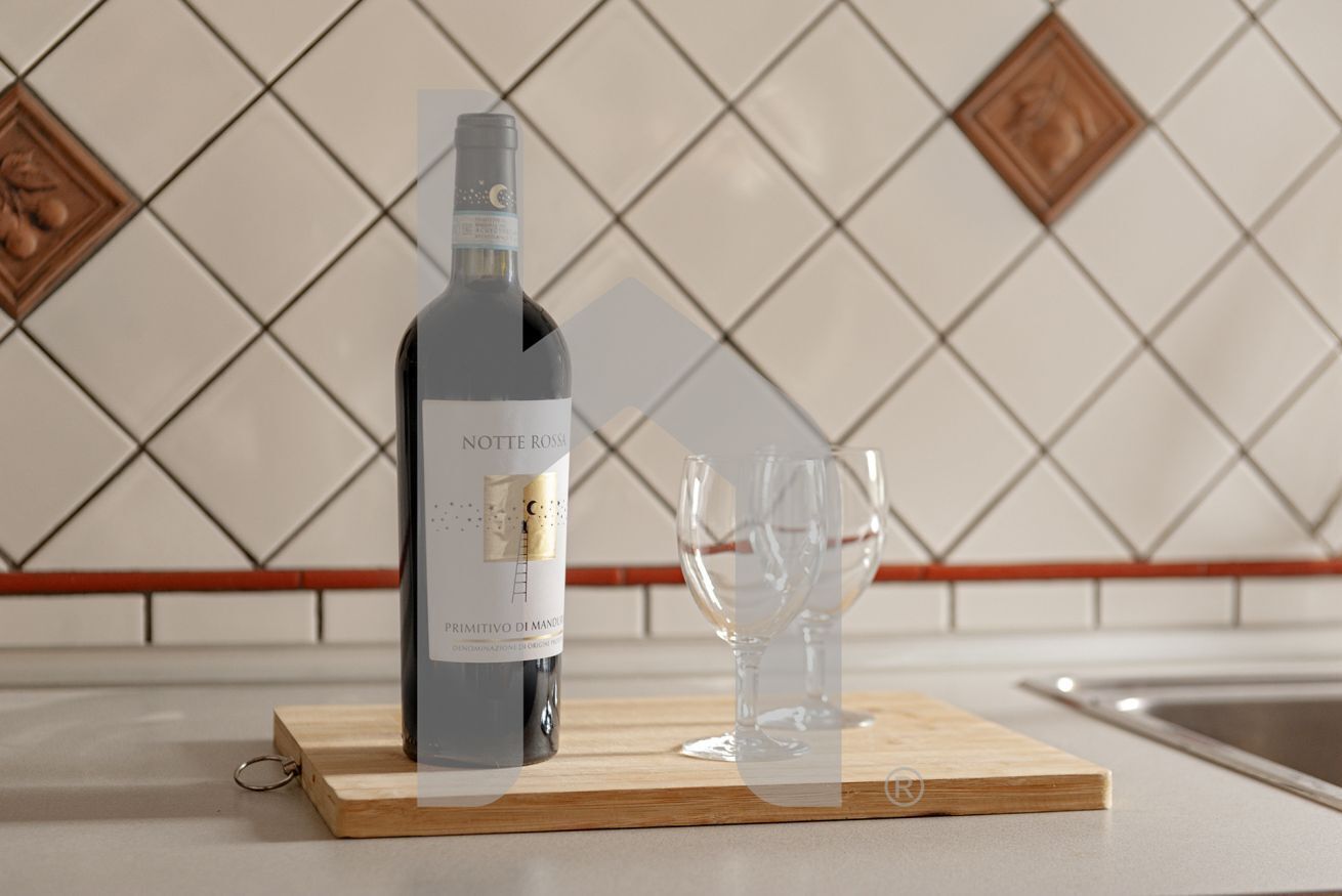 Une bouteille de vin posée sur une planche en bois, avec deux verres à côté, le tout sur un fond de carreaux.