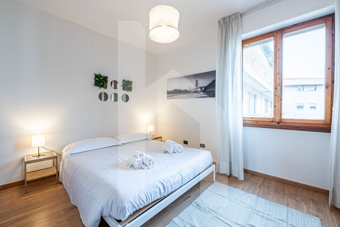 A modern bedroom with a bed, bedside lamps, a window with curtains, and a wall-mounted picture.