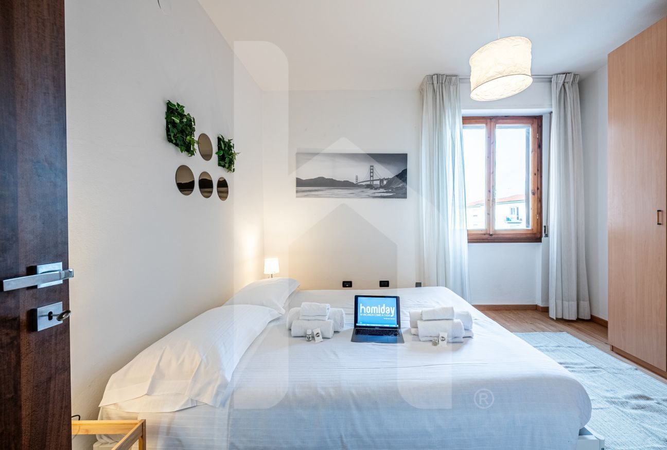 A hotel room with a bed, a laptop displaying 'homiday', a window with curtains, and a painting of a bridge.