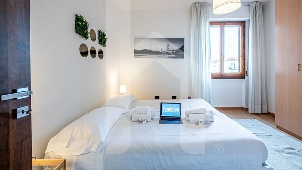 A hotel room with a bed, a laptop displaying 'homiday', a window with curtains, and a painting of a bridge. - [Homiday] Elegant apartment with wooden finishes in the central area.Relax and enjoy the three bright double bedrooms equipped with air conditioning and heating. Homiday offers the perfect accommodation for those who travel for work or leisure, in the ar (Pisa)