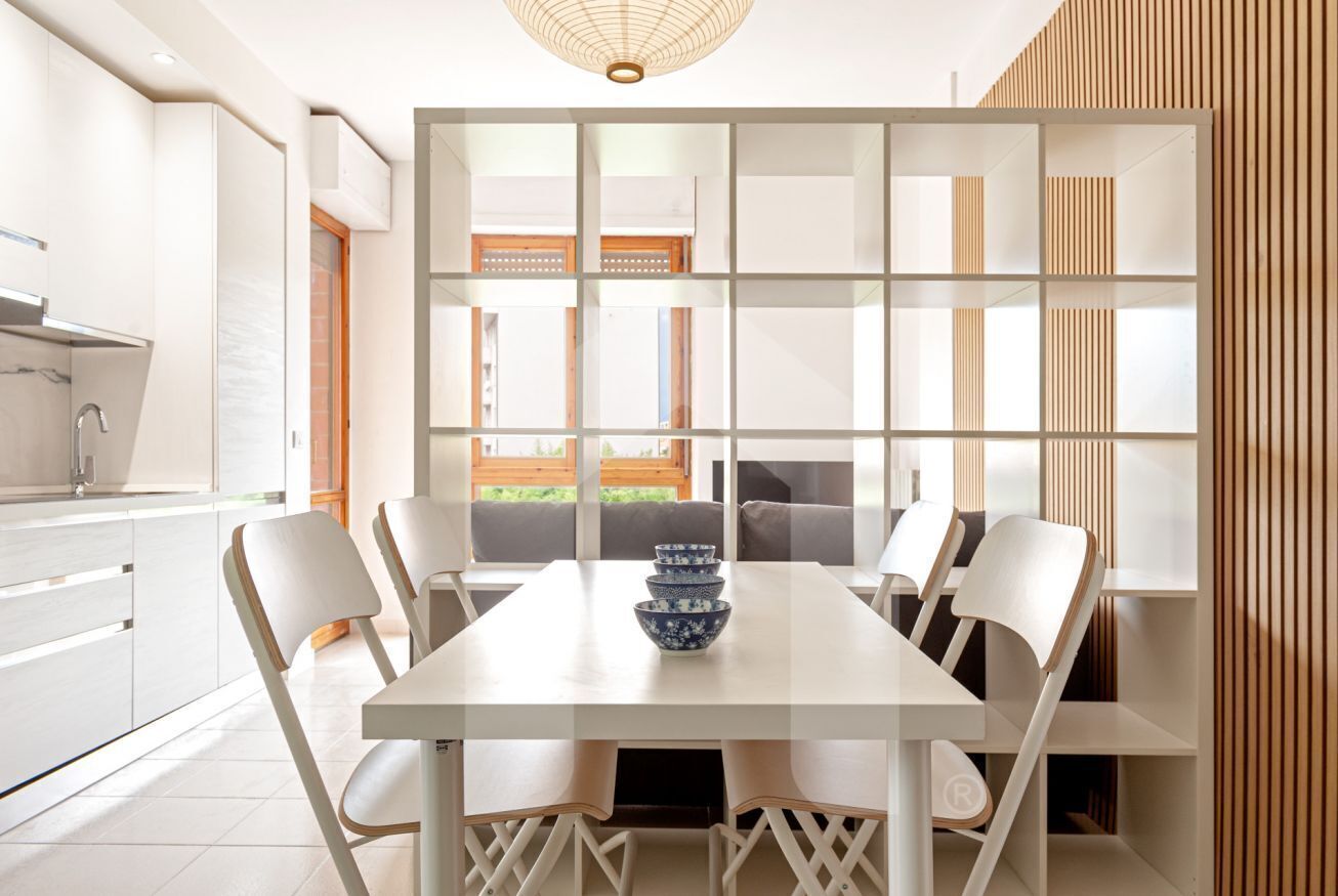 A modern kitchen with white cabinetry, a dining table with chairs, and a view through a partition to the outside.