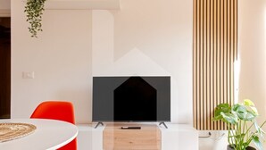 Ein modernes Wohnzimmer mit einem Flachbildfernseher auf einem Standfuß, einem Holztisch, einem leuchtend orangefarbenen Sessel und einem Blumentopf.