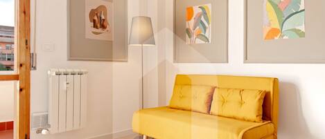 A modern living room with a yellow sofa, framed artwork on the walls, a floor lamp, and a radiator.