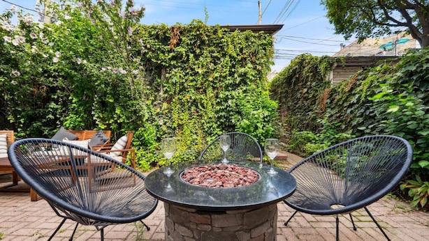 An outdoor seating area with a stone fire pit table surrounded by two black wire chairs, set against a backdrop of lush greenery and a brick pathway. - Wrigleyville 2BR w/Parking, Firepit & Workspace for Remote Work (Chicago)