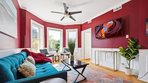 A living room with a teal sofa, a coffee table, and a colorful rug. There are windows, a ceiling fan, and a TV mounted on the wall. - Wrigleyville 2BR w/Parking, Firepit & Workspace for Remote Work (Chicago)