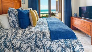 Een slaapkamer met een groot bed, een houten hoofdbord, een plafondventilator, een televisie op een houten kast en een balkon met uitzicht op de oceaan.
