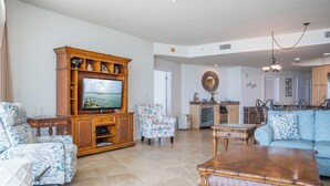 A spacious living room with a large flat-screen TV, a wooden entertainment unit, patterned armchairs, a blue sofa, and a herringbone-patterned coffee table.