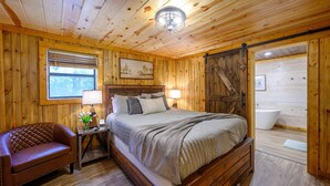 Un dormitorio tipo cabaña de madera con una cama, una mesita de noche, un sillón de cuero, una ventana y un baño visible a través de una puerta abierta.