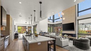 A modern kitchen with a central island, a dining area with a sofa and chairs, a fireplace, and large windows offering a view of the city. - Abode at Moose Ridge, private hot tub, ski access, modern home (Park City)