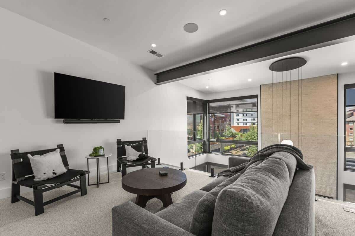 Una sala de estar moderna con un sofá, dos sillones, una mesa de centro redonda, un televisor de pantalla plana montado en la pared y ventanas amplias con vistas a edificios.