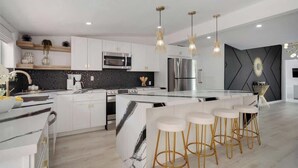 A modern kitchen with white cabinets, a marble island, and gold bar stools. - Chic Palm Escape w/ Private Pool (Palm Desert)